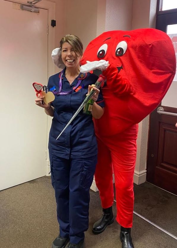 A female nurse stands in front of a red heart mascot whilst holding a rose, chocolate heart and bottle of wine